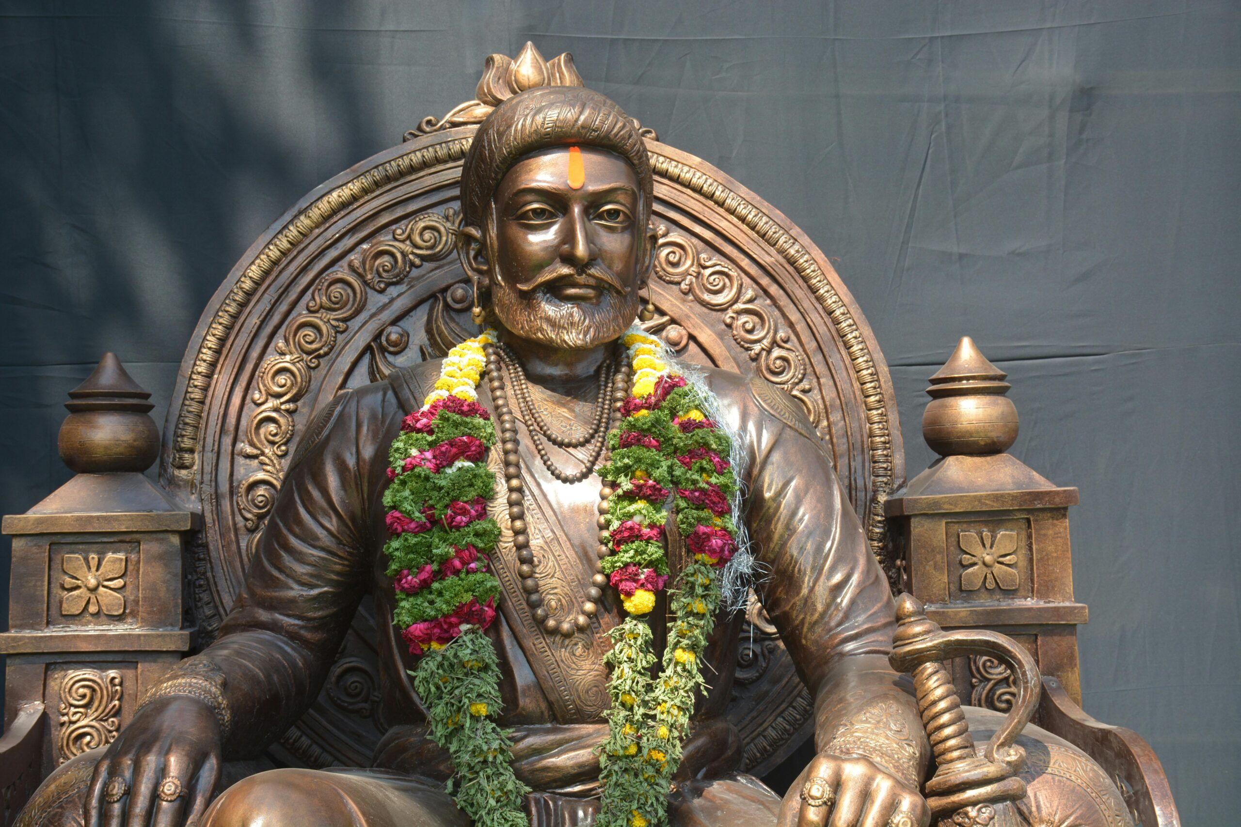 Bronze statue of Shivaji Maharaj adorned with vibrant floral garland under daylight.